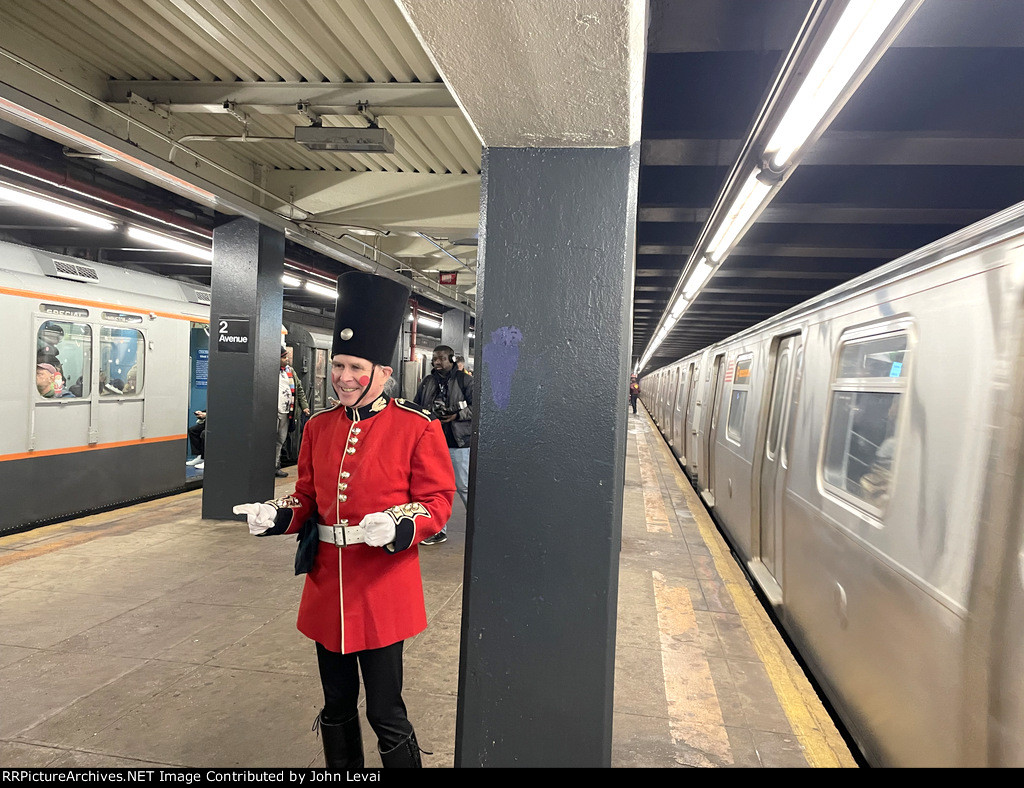 A man wearing a British soldier outfit at 2nd Ave Station walking on the platform between the R179 consist F train and the Arnine consist making up the Holiday Train